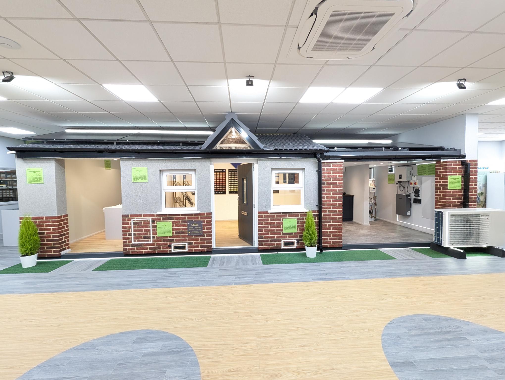 Full-size model house inside the Infinity Energy Organisation Wembley showroom, with cutaway walls showing external and internal wall insulation, a tiled roof, and a Daikin air source heat pump unit alongside