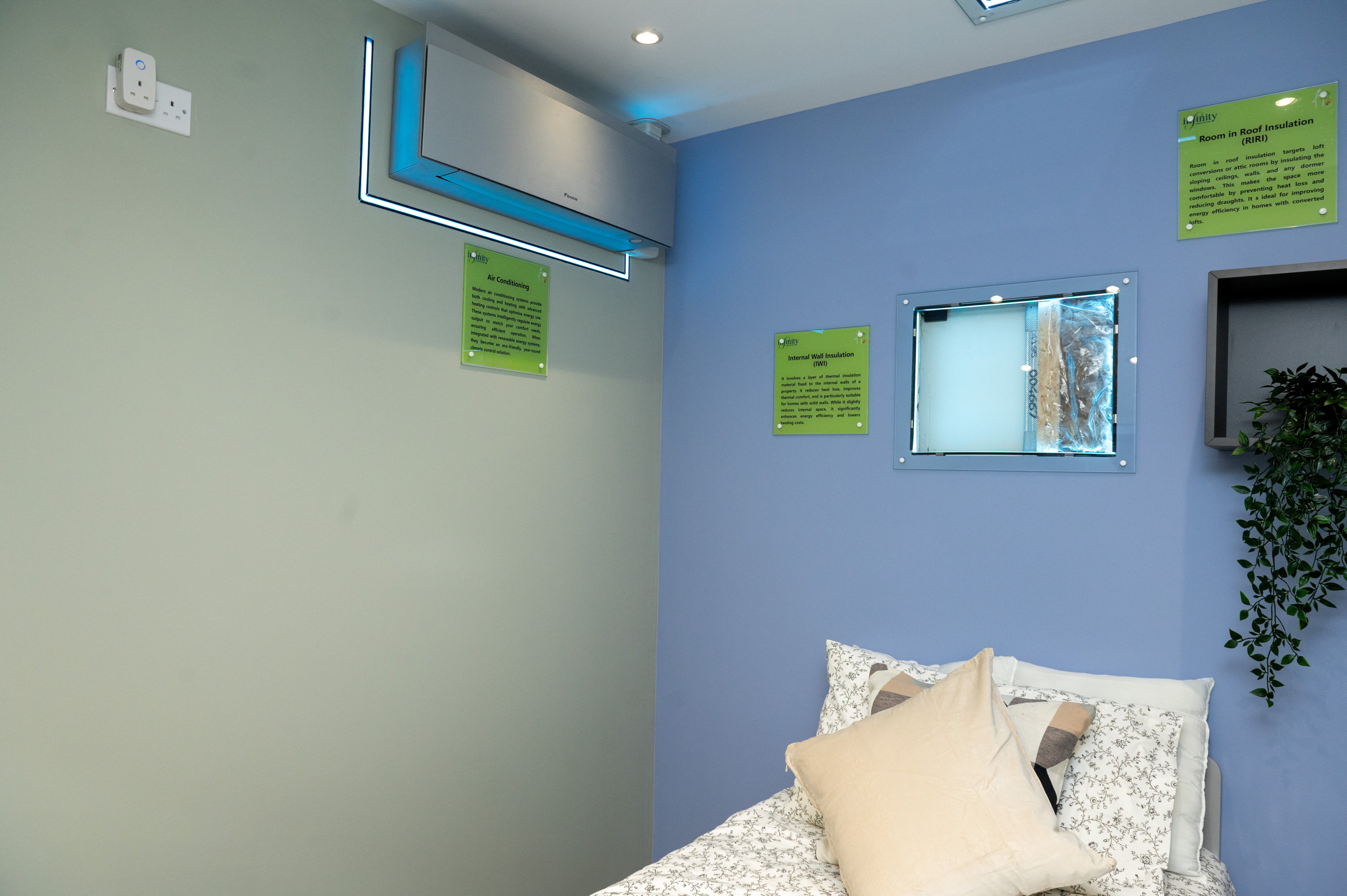 Showroom bedroom display with a wall-mounted air conditioning unit and cutaway panels demonstrating internal wall insulation and room-in-roof insulation
